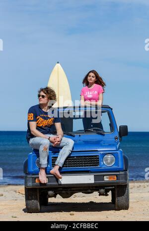 Junge, energische Frau mit Surfbrett und attraktivem Mann, der an hellem Tag auf blauem Offroad-Spieler sitzt Stockfoto