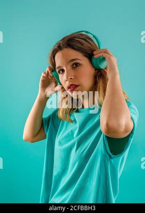 Seitenansicht einer nachdenklichen jungen Frau in einem hellen T-Shirt, die auf die Kamera schaut, während sie auf türkisfarbenem Hintergrund Musik in Kopfhörern hört Stockfoto