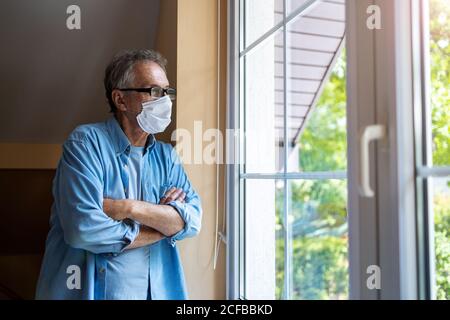 Älterer Mann mit schützender Gesichtsmaske, der aus dem Fenster schaut Stockfoto
