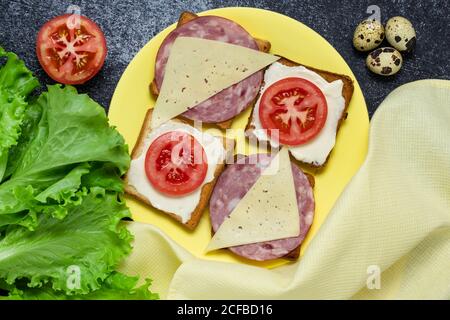 Sandwiches mit Tomaten, Wurst und Käse auf einem gelben Teller. Salatblätter und Wachteleier. Frühstückskonzept, Stillleben. Toast zum Mittagessen. Flat la Stockfoto