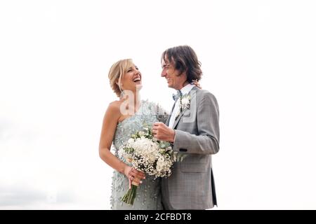 Zufrieden jungen Bräutigam im Hochzeitsanzug stolz Blick auf schöne Blonde behaarte Braut in stilvollem Kleid hinter bei leerem Sand Meeresküste Stockfoto