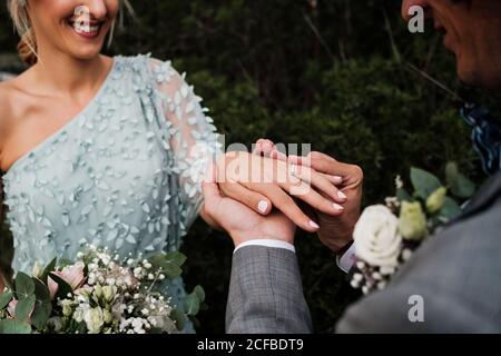 Von oben vom Bräutigam im Hochzeitsanzug Ring an Finger der glücklichen zukünftigen Frau mit grünen Pflanzen auf dem Hintergrund Stockfoto