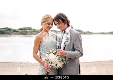 Zufrieden jungen Bräutigam im Hochzeitsanzug stolz Blick auf schöne Blonde behaarte Braut in stilvollem Kleid hinter bei leerem Sand Meeresküste Stockfoto