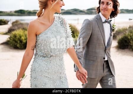 Zufrieden jungen Bräutigam im Hochzeitsanzug stolz Blick auf schöne Blonde behaarte Braut in stilvollem Kleid hinter bei leerem Sand Meeresküste Stockfoto