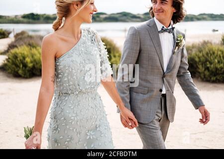 Zufrieden jungen Bräutigam im Hochzeitsanzug stolz Blick auf schöne Blonde behaarte Braut in stilvollem Kleid hinter bei leerem Sand Meeresküste Stockfoto