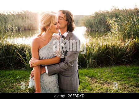 Stilvolles frisch verheiratetes Paar in Hochzeitsanzügen umarmen und küssen Mit grünen Pflanzen und nur an sonnigen Tag Stockfoto