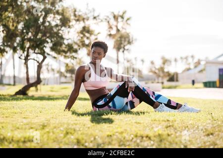 Schlanke afroamerikanische Athletin in bunten activewear und weiß Sneaker blicken neugierig weg, während sie auf grünem Gras sitzen Auf Rasen und Ruhe nach dem Training Stockfoto