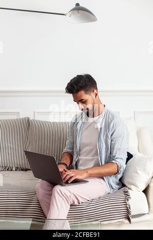 Junge ethnische Mann in lässigen Kleidern sitzen auf Couch und Browsing Laptop im Wohnzimmer der modernen Wohnung Stockfoto