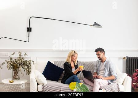 Positive blonde Frau mit einer Tasse Saft, die im Wohnzimmer der modernen Wohnung auf dem Smartphone surft und auf der Couch in der Nähe eines ethnischen Freundes sitzt, der auf der Laptop-Tastatur tippt Stockfoto