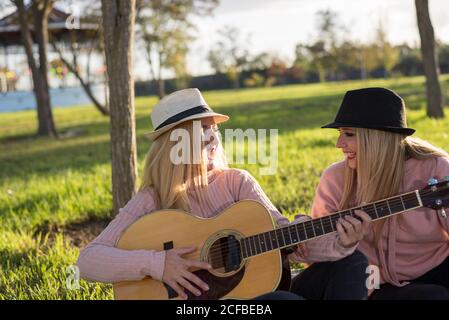 Kaukasische blonde Mädchen in Hüten. Glücklich singen und Gitarre spielen. Sitzen auf einer Decke im Park. Stockfoto