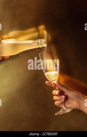 Anonyme Ernte Person hält Weinglas, während eine andere gesichtslose Person Gießen Champagner in Glas aus der Flasche während der Party auf goldenem Hintergrund Stockfoto
