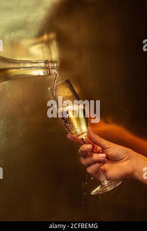 Anonyme Ernte Person hält Weinglas, während eine andere gesichtslose Person Gießen Champagner in Glas aus der Flasche während der Party auf goldenem Hintergrund Stockfoto