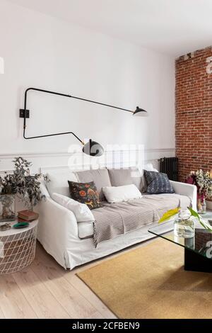 Bequemes Sofa mit weichen Kissen unter moderner Metalllampe Neben Tischen mit Dekorationen in stilvollen gemütlichen Wohnzimmer an Zu Hause Stockfoto