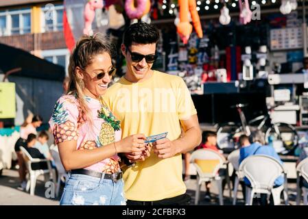 Aufgeregte Frau in Sonnenbrillen und legerer Kleidung, die Lotterielos ansieht, mit einem hübschen Mann, der die Zeit auf dem Messegelände genießt Stockfoto