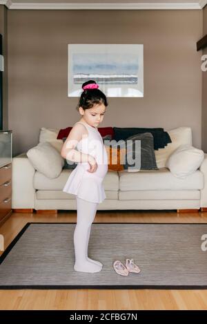 Nettes kleines Mädchen in hellrosa Trikot und Strumpfhosen Putting Auf Rock beim Stehen in der Nähe von Tanzschuhen in gemütlichen Wohn Zimmer zu Hause Stockfoto