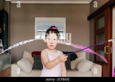 Fokussierte niedliche kleine Brünette Mädchen in Trikot Blick auf Kamera Beim Spinning Band während rhythmischer Gymnastik üben Training in cosy Wohnzimmer zu Hause Stockfoto