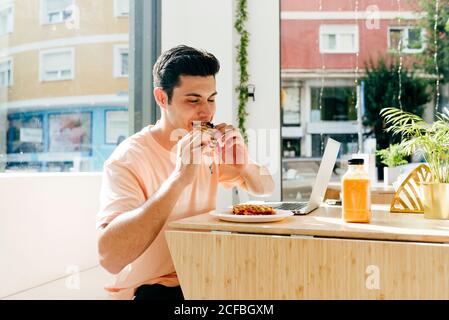 Glücklicher junger Mann mit geschlossenen Augen beißen süßen Snack während Sitzen am Tisch mit Laptop gegen Fenster im Restaurant Stockfoto