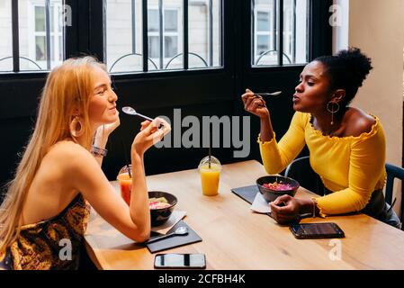 Seitenansicht einer jungen blonden Frau und einer afroamerikanischen Freundin, die mit Tassen Saft am Tisch sitzt und köstliche Smoothie-Schüsseln genießt, während sie im Café frühstücken Stockfoto