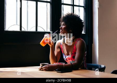 Seitenansicht der eleganten afroamerikanischen jungen Frau mit Kopfhörern Am Hals frühstücken und Fruchtsaft trinken, während man sitzt Am Tisch neben dem Fenster und genießen Sie den Morgen im Café Stockfoto