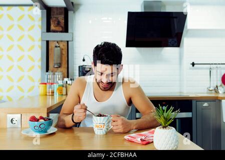 Junge lächelnd schöner Mann mit stilvoller Frisur in lässigen ärmellosen hemd hält Tasse Kaffee mit Schokolade in der Küche Stockfoto