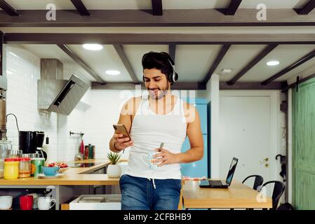 Junger, fröhlicher, erfolgreicher Mann mit Kopfhörern, der auf einem Stuhl sitzt, Smartphone stöbert und in der Küche eine Tasse heißen Kaffee hält Stockfoto