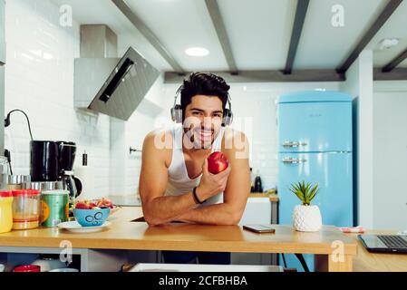 Junge lächelnd schöner Mann mit stilvollen Frisur in Headset lehnte Auf dem Tisch in der Küche und hält reifen Apfel Stockfoto