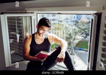 Seitenansicht von jungen ernst schönen Mann in lässig ärmellos hemd sitzt am Fenster und liest Interessantes Buch Stockfoto