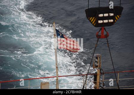 Blick auf fliegende liberianische Flagge, Hecklicht und gesicherter Kranhaken in der Achterstation des Frachtschiffes unterwegs durch den ruhigen Ozean im Sommer. Stockfoto