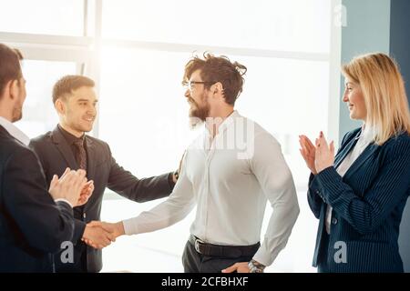 Zwei fröhliche Geschäftsleute schüttelten sich die Hände, während ihre Kollegen applaudierenden Und lächelnd im Hintergrund Stockfoto