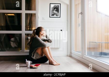Seitenansicht der ruhigen jungen Frau in Freizeitkleidung suchen Am Fenster nachdenklich sitzen auf dem Boden mit einer Tasse Kaffee und Toast während des Frühstücks zu Hause Stockfoto
