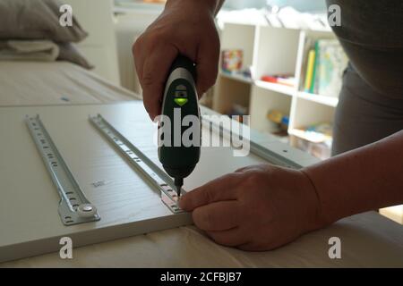 Ein Mann, der für seinen Sohn in seinem Zimmer eine Schublade eines Computerschreibtisches mit einem Akkuschrauber aus der Nähe zusammengebaut hat. Er benutzt ein Werkzeug und eine Anleitung. Stockfoto