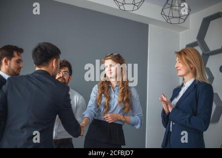 Zwei fröhliche Geschäftsleute schüttelten sich die Hände, während ihre Kollegen applaudierenden Und lächelnd im Hintergrund Stockfoto
