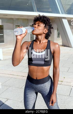 Junge fröhliche afroamerikanische Läuferin in Sport top und Leggings stehen neben Gebäude und Trinkwasser während mit Pause während des Trainings in der Stadt Stockfoto