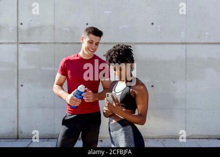 Fröhlicher Mann mit einer Flasche Wasser und eine afroamerikanische Frau, die sich das Smartphone teilt, stehen zusammen gegen eine Betonwand und nutzen eine App für das Training während einer Pause in der Stadt Stockfoto