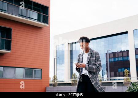 Von unten von glücklichen jungen afroamerikanischen männlichen Studenten in Stylisches Outfit beim Surfen auf dem Smartphone, während Sie auf der Stadtstraße in der Nähe stehen Modernes Gebäude Stockfoto