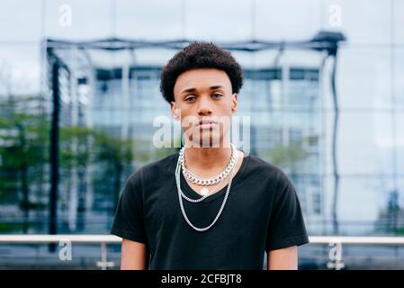 Von unten von jungen lockig behaarten afroamerikanischen Teenager-Kerl In schwarz T-Shirt und trendigen Nackenketten Blick auf Kamera, während Sie gegen ein verschwommenes modernes Gebäude mit Glaswand stehen Stockfoto