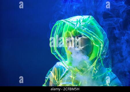Junge ziemlich ungewöhnliche asiatische Frau in Kunststoff transparent Regenmantel und gelbes Haar Rauchen in fluoreszierendem Licht Stockfoto
