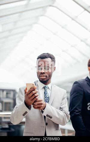 Afroamerican bärtigen Geschäftsmann tragen Anzug und Brille SMS auf dem Telefon in der Mall oder Halle der moderne Büro. Business Konzept Stockfoto