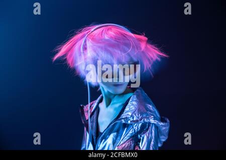 Junge subkulturelle asiatische Frau mit rosa verstreuten Haaren und silberner Jacke tanzt, während sie Musik in Kopfhörern hört Stockfoto