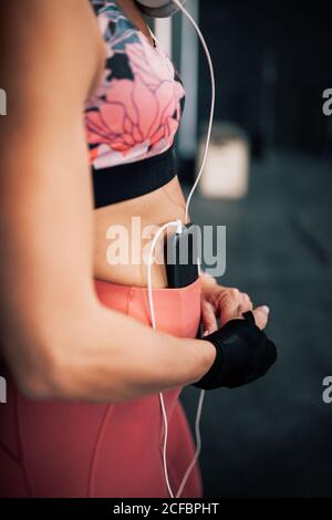 Seitenansicht der Crop Boxerin in Sportbekleidung mit Verband Am Handgelenk Musik hören mit Smartphone und Kopfhörer während Ruhe nach dem Training im Fitnessstudio Stockfoto