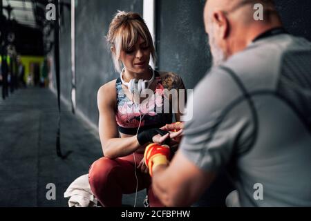 Erwachsener bärtiger männlicher Ausbilder, der Frau Sportlerin dabei hilft, einen Verband am Handgelenk zu wickeln, während sie in der Nähe der schwarzen Wand sitzt und sich für das Training im Boxstudio vorbereitet Stockfoto