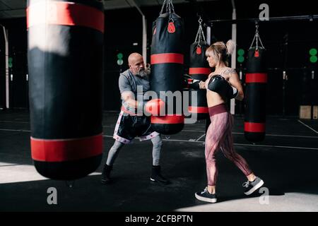 Seitenansicht der selbstbewussten jungen Boxerin in Boxhandschuhen Schlagen schweren Boxsack während des Trainings mit männlichen Lehrer in Moderner Fitnessraum Stockfoto