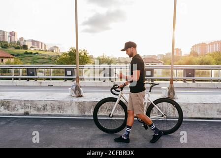 Seitenansicht des jungen beiläufigen bärtigen Radfahrers in schwarz Cap mit Telefon beim Gehen mit dem Fahrrad auf der Straße mit Stadtgebäude im Hintergrund Stockfoto