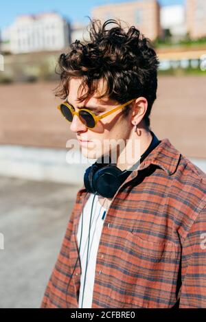 Hübscher junger Mann in kariertem Hemd und Sonnenbrille beim Betrachen Kamera auf unscharfem Hintergrund der Stadtstraße stehend Stockfoto