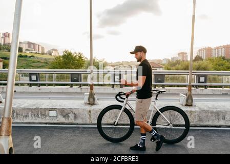 Seitenansicht des jungen beiläufigen bärtigen Radfahrers in schwarz Cap mit Telefon beim Gehen mit dem Fahrrad auf der Straße mit Stadtgebäude im Hintergrund Stockfoto