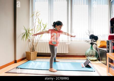 Seitenansicht des positiven kleinen Mädchens, das in Yoga-Pose steht Auf der Matte neben dem Fenster zu Hause beim Yoga üben und Video-Tutorials auf dem Tablet ansehen Stockfoto