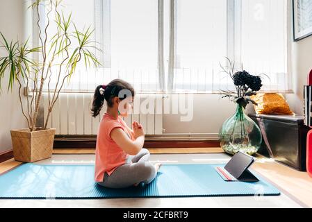 Kleines Mädchen, das in Lotuspose sitzt und auf einer Matte meditiert Mit geschlossenen Augen zu Hause mit großem Fenster beim Üben Yoga und Video-Tutorials auf dem Tablet Stockfoto