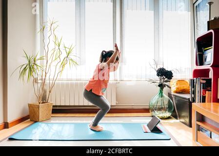 Seitenansicht des positiven kleinen Mädchens, das in Yoga-Pose steht Auf der Matte neben dem Fenster zu Hause beim Yoga üben und Video-Tutorials auf dem Tablet ansehen Stockfoto