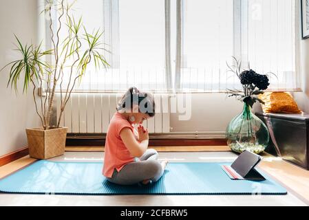 Kleines Mädchen, das in Lotuspose sitzt und auf einer Matte meditiert Mit geschlossenen Augen zu Hause mit großem Fenster beim Üben Yoga und Video-Tutorials auf dem Tablet Stockfoto