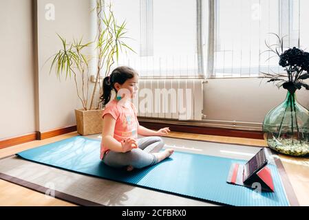 Kleines Mädchen, das in Lotuspose sitzt und auf einer Matte meditiert Mit geschlossenen Augen zu Hause mit großem Fenster beim Üben Yoga und Video-Tutorials auf dem Tablet Stockfoto